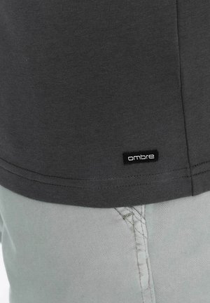 Graues Baumwoll-T-Shirt mit abgerundetem Saum, das ein kleines schwarzes "Ombre"-Etikett nahe dem unteren Saum zeigt. Kombiniert mit hellgrünen Hosen.