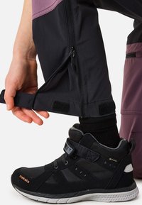 Sorte og lilla bukser med lynlås og velcrolukninger ved manchetterne, kombineret med sorte høje sko med mesh- og teksturerede materialer samt reflekterende detaljer.