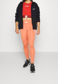Zwarte hoodie met ritssluiting, rood cropped shirt met Nike-logo, perzikkleurige legging met zijkanttekstpatroon en zwarte sportschoenen.