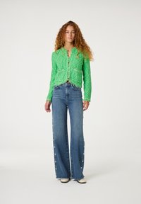Groene gestructureerde jas met rafelige randen, gecombineerd met hoge taille blauwe flared jeans met hartvormige knopen langs de zijkanten.