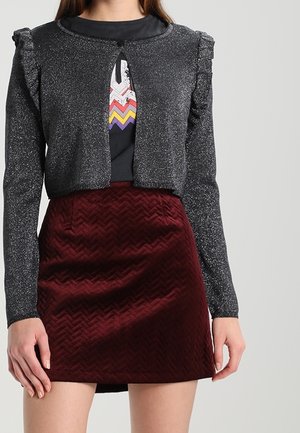 Kobieta ubrana w błyszczący czarny krótki sweter z długim rękawem, czarny top z grafiką oraz fakturowaną ciemnoczerwoną welwetową mini spódnicę.