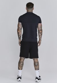 Melns īs piedurkņu krekls, kas saskaņots ar melnām šortiem, ar rakstainiem tetovējumiem uz kājām, baltām sporta zeķēm un melnām kedām.