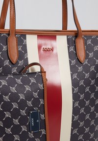Nákupní taška z tmavě modrého texturovaného materiálu s bílým vzorem, hnědými koženými uchy a středovým červeno-krémovým pruhem. Zobrazené logo značky.
