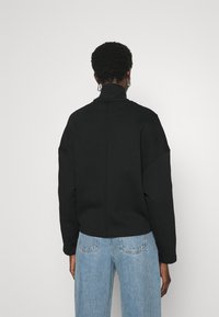 Giacca nera a pullover con colletto alto, spalle scese e vestibilità morbida. Abbinata a jeans in denim azzurro chiaro. Texture liscia.