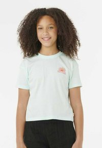 Chica sonriente con cabello rizado, vistiendo una camiseta de Ripcurl color azul claro y pantalones negros, de pie frente a un fondo blanco liso.