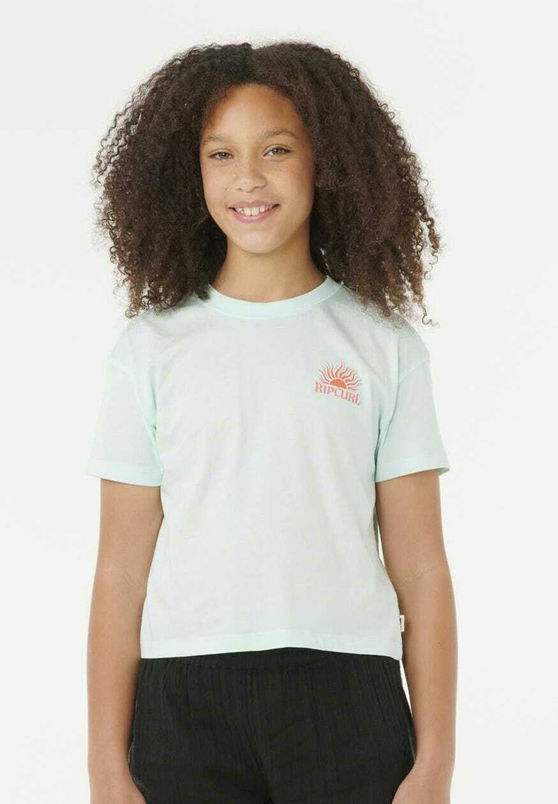 Chica sonriente con cabello rizado, vistiendo una camiseta de Ripcurl color azul claro y pantalones negros, de pie frente a un fondo blanco liso.