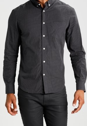 Uomo con camicia a quadri grigia scura con taschino e pantaloni neri, in piedi davanti a uno sfondo semplice.