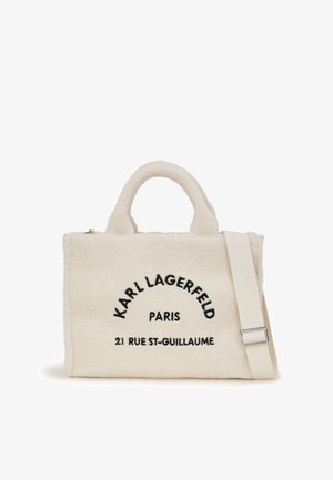 Kabelka z krémového umělého ovčího rouna s černě vyšitým názvem značky a adresou. Má dvojité horní uchy a odnímatelný ramenní popruh.