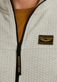 Veste zippée texturée gris clair avec une fermeture éclair noire, arborant un logo PME Legend jaune et noir sur la poitrine, portée sur une chemise moutarde.