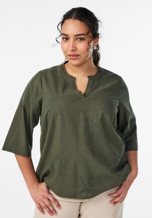 Kähara juustega naine, kes kannab tumerohelist tippu lillemustriga ja beeže pükse, seisab käed puusadel.