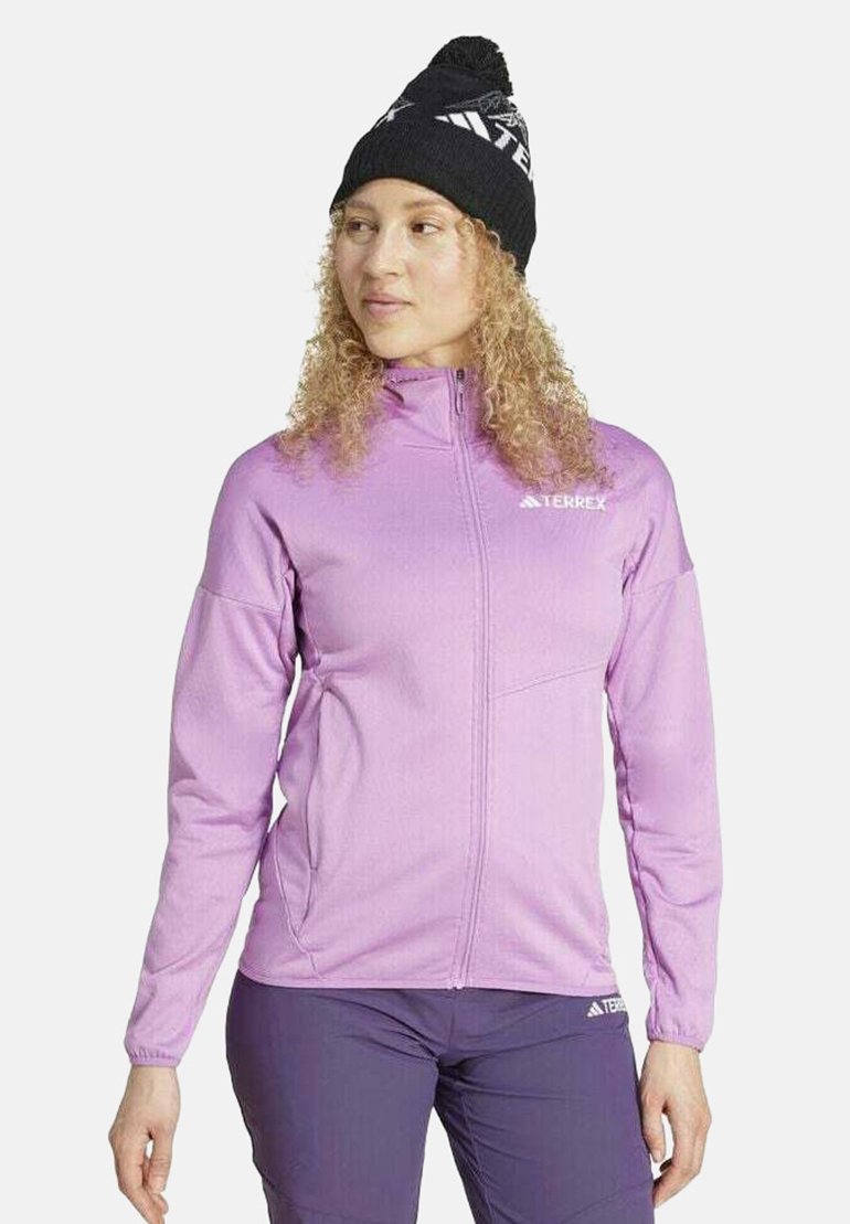 Donna che indossa un berretto nero con pon pon, giacca viola chiaro con cerniera e logo Terrex, e pantaloni viola scuro, in piedi davanti a uno sfondo bianco.