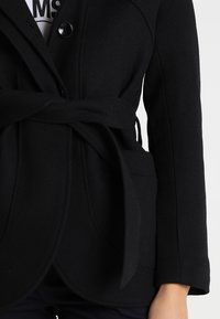 Blazer negro hecho de un tejido texturizado, con un cinturón ajustado en la cintura, cierre de botón único y dos bolsillos frontales.