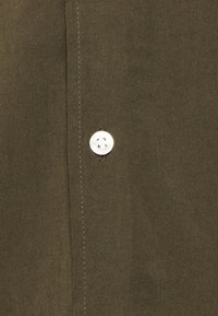 Tissu vert olive avec une texture lisse, présentant un bouton blanc et une couture contrastante le long du bord. Bouton situé près de la couture.