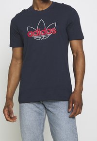 Marinblå bomulls-T-shirt med en röd och vit adidas-logotyp med bladdesign, korta ärmar och rund halsringning.