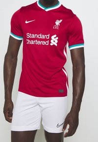Camiseta de fútbol roja de Nike con acentos blancos, con cuello en V y mangas cortas. Logo de "Standard Chartered" impreso en blanco. Pantalones cortos blancos debajo.