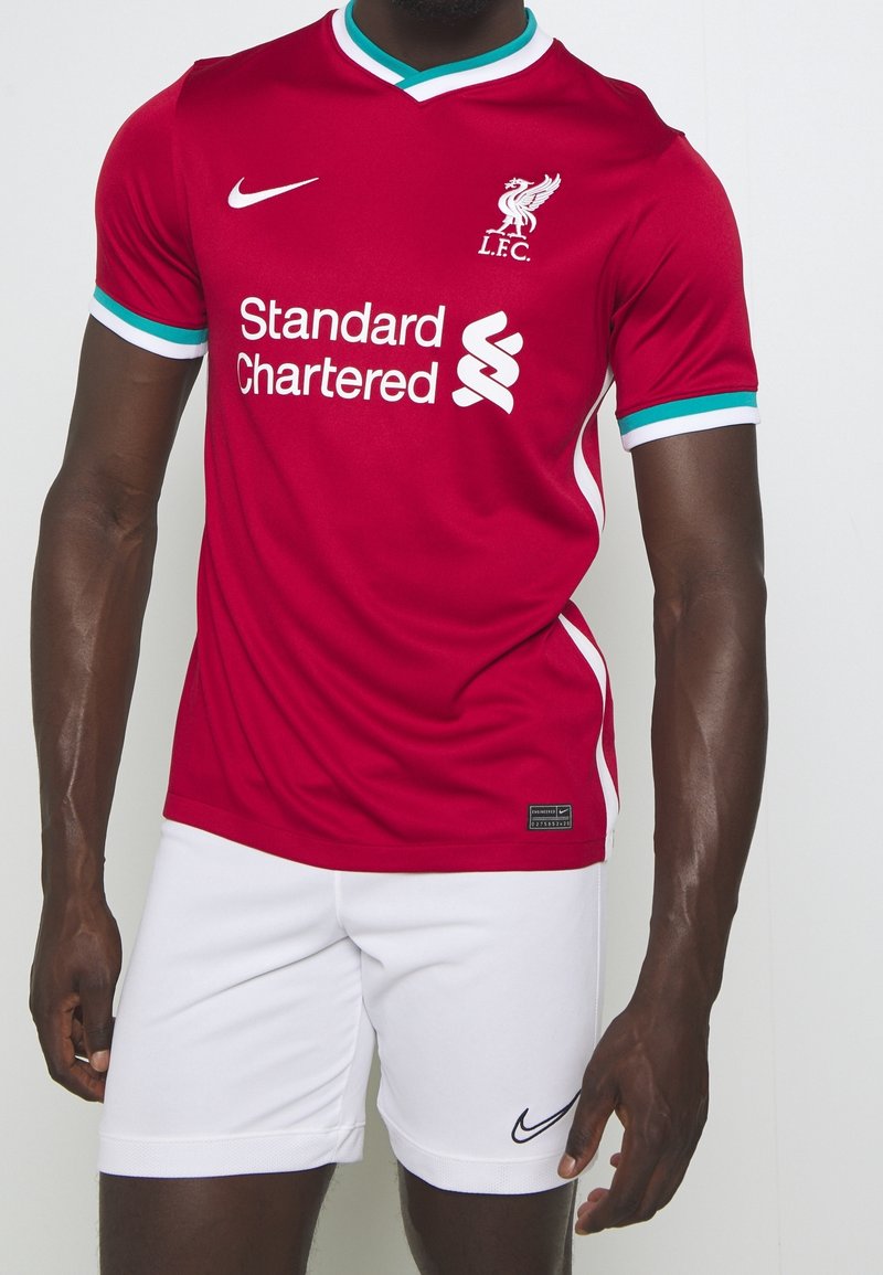 Camiseta de fútbol roja de Nike con acentos blancos, con cuello en V y mangas cortas. Logo de "Standard Chartered" impreso en blanco. Pantalones cortos blancos debajo.