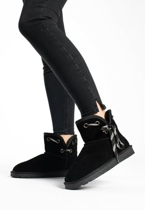 Botas de tobillo de ante negro con puntera redondeada, cordones de patrón gris y blanco, y ojales decorativos. La caña está ligeramente forrada de pelo.