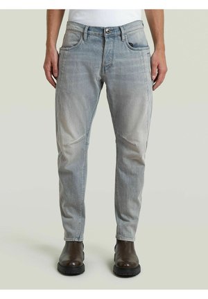 Persona che indossa jeans grigi slavati e stivali di pelle marrone scuro in piedi davanti a uno sfondo chiaro e uniforme.