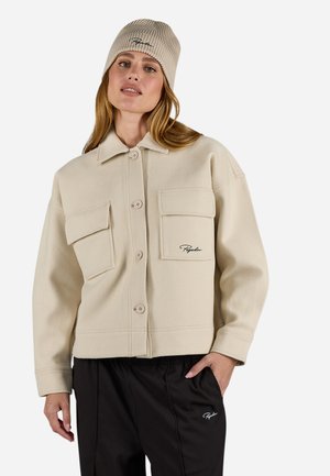 Beige fleece jacket with two front pockets, button closure, and embroidered logo. Paired with black pants and a ribbed beige beanie.