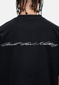 Black cotton t-shirt with a crew neck. Features white cursive text "Good For Nothing" embroidered on the back. Smooth texture, relaxed fit.