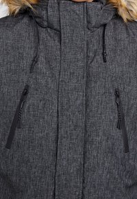 Botón gris oscuro acolchado con una tela texturizada, que cuenta con una capucha forrada de piel sintética, cordones y dos bolsillos laterales con cremallera.