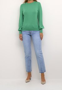 Femme portant un haut vert à manches longues, un jean droit bleu clair et des talons pointus beige, debout devant un fond uni.