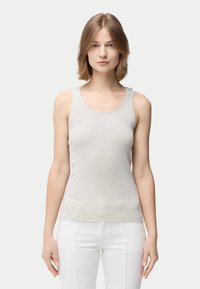 Ung kvinna med axellångt ljusbrunt hår, iklädd en ljusgrå ärmlös ribbad topp och vita byxor, stående mot en enfärgad bakgrund.