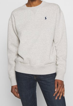 Jasnoszara bluza crewneck z długimi rękawami i granatowym logo Polo Ralph Lauren na lewej piersi, noszona z ciemnoniebieskimi dżinsami.