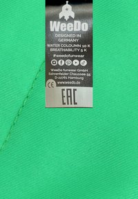 Tessuto verde con una superficie texturizzata, caratterizzato da una cucitura. Un'etichetta con testo nero che mostra i dettagli del marchio e le specifiche di resistenza all'acqua.