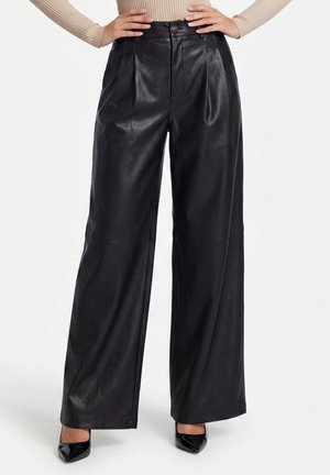 Woman wearing high-waisted black wide-leg leather trousers with pleats, a beige ribbed long-sleeve top, and black pointed-toe heels.
