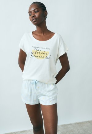 Vrouw die een wit shirt draagt met de tekst "Als het leven je citroenen geeft, maak dan limonade!" en lichtblauwe gestreepte korte broek met kleine gele patronen.