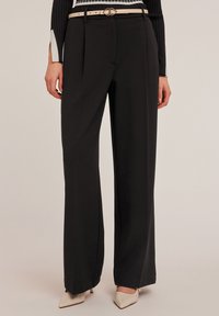 Zwarte wijde broek van gladde stof, met een ceintuur in de taille en een lichte ceintuur, gecombineerd met puntige beige hakken.