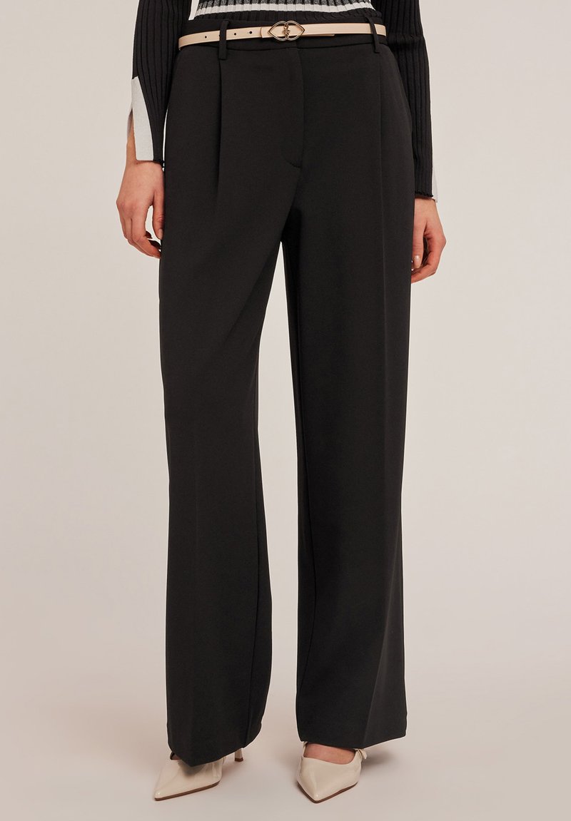 Zwarte wijde broek van gladde stof, met een ceintuur in de taille en een lichte ceintuur, gecombineerd met puntige beige hakken.