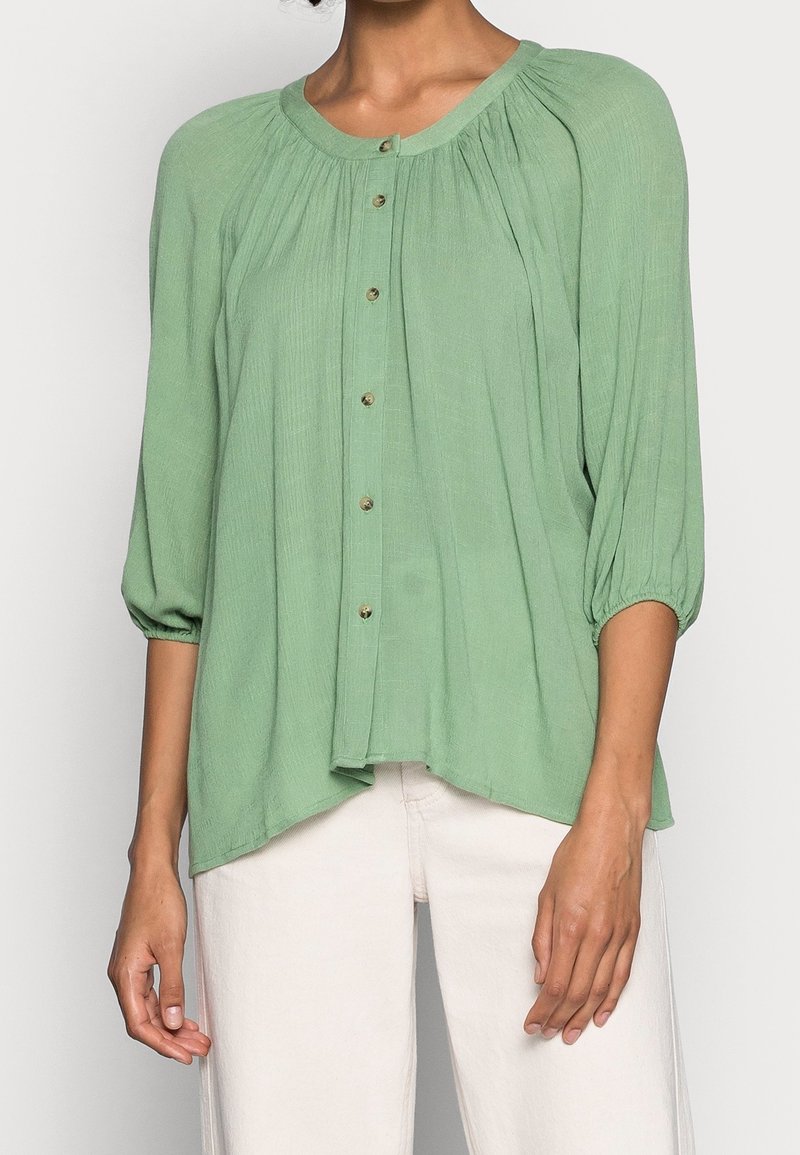 Blouse boutonnée vert clair à manches trois-quarts plissées, portée avec un pantalon blanc cassé sur une personne debout.