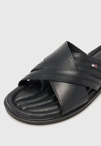 Sandale glissante en cuir noir avec un design de brides entrecroisées, semelle rembourrée et texture lisse avec un détail de marque minimaliste.