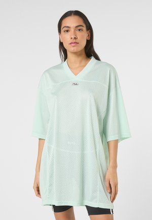 Giovane donna con lunghi capelli scuri, indossa una maglia sportiva oversize FILA color verde menta chiaro traforata e pantaloncini neri, in piedi.