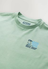 Lysegrøn bomulds t-shirt med et tegneseriegrafik og teksten "HIGH TIDE SURF SHOP" i blå og sort. Løs pasform og rund hals.