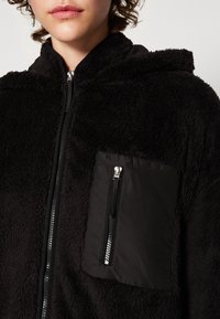 Chaqueta de forro polar negra con una apariencia suave y texturizada; presenta un bolsillo con cremallera en la parte inferior delantera y una capucha estándar.