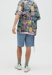 Personne portant une chemise à imprimé abstrait coloré, un short en jean bleu, des chaussettes grises et des baskets blanches, debout de dos face au spectateur.