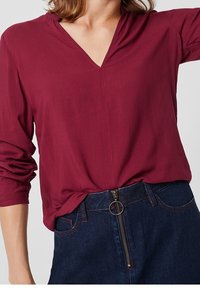 Bordeauxfärgad långärmad blus med V-ringning och subtil sömnad. Bärs med en mörk denimkjol som har en synlig dragkedja och ringdragning.