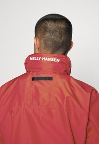 Piros vízálló dzseki magas gallérral, hátul fehér "Helly Hansen" logóval. Simma textúra, minimalista dizájn.