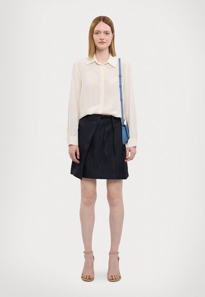 Lys kremfarget skjorte med knapper, marineblå plisserte skjørt med knytedetalj, og beige sandaler med stropper. En liten blå crossover-veske fullfører antrekket.