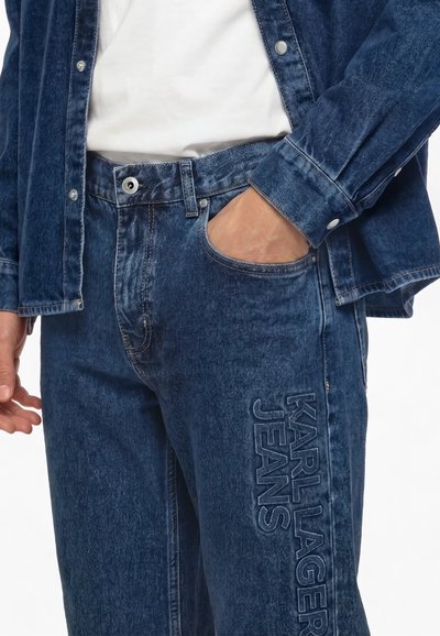 Personne portant un jean en denim bleu foncé et une veste, avec une main dans la poche et "KARL LAGERFELD JEANS" brodé sur la cuisse.