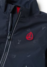 Cazadora de viento navy con forro rojo, que presenta un prominente parche de logo rojo y sutiles patrones de velero. Cremallera con costuras en contraste.