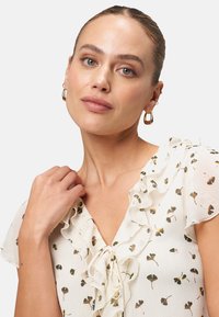 White blouse with ruffled neckline, featuring a floral pattern in green and gold. Smooth fabric texture and short, flutter sleeves.