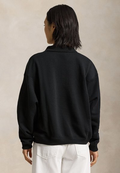 Vállig érő sötét hajú személy, aki laza fekete pulóvert és fehér nadrágot visel, háttal egy bézs háttér előtt.