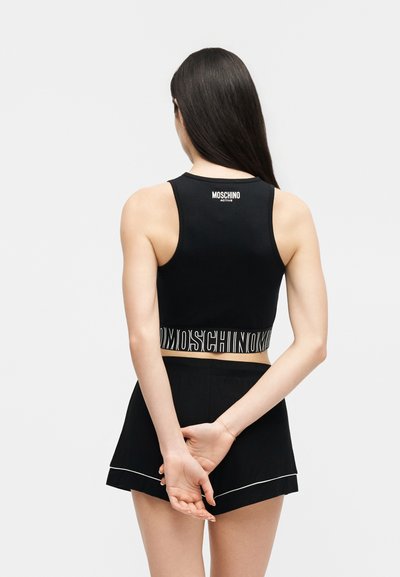 Žena s dlouhými tmavými vlasy, oblečená v černém krátkém topu Moschino Active a černých šortkách, stojící s rukama sepnutýma za zády.