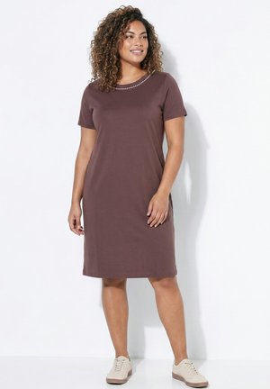 Frau mit lockigem Haar, die ein schlichtes braunes kurzärmeliges Kleid und weiße Sneakers trägt, vor einem weißen Hintergrund stehend.
