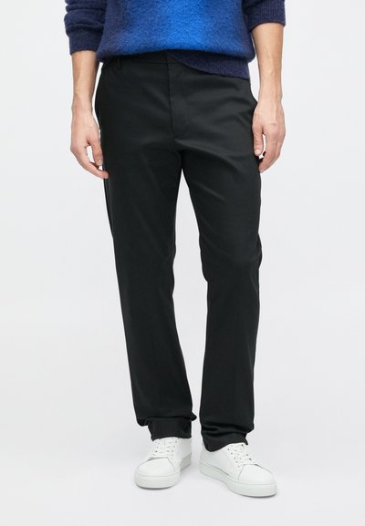 Black tapered pants made of smooth fabric, featuring side pockets and a tailored fit, paired with white sneakers on a neutral background.