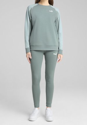 Person iført grøn Puma træningsdragt med sweatshirt og leggings, kombineret med hvide sneakers, stående mod en ensfarvet baggrund.